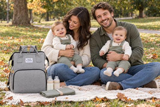 Mom and dad holding their twins at an autumn park picnic, with a gray diaper bag backpack, bottles, and a changing mat beside them.